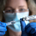 COVID Vaccine Sales CRASH Person with gloves holding COVID-19 vaccine syringe.