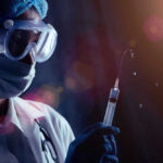 Person in PPE holding syringe with liquid droplets.