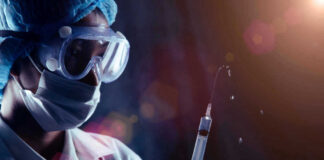 Person in PPE holding syringe with liquid droplets.