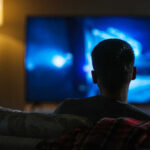 Person sitting on a couch watching a television screen in a dimly lit room