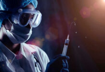 Person in PPE holding syringe with liquid droplets.
