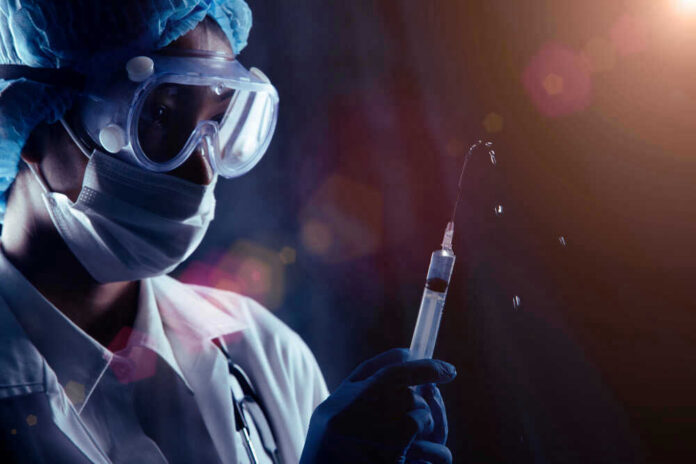 Person in PPE holding syringe with liquid droplets.