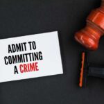 A hand holding a card that says 'ADMIT TO COMMITTING A CRIME' next to a gavel and a shaving razor