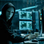 Person in hoodie and mask using computer, multiple screens.