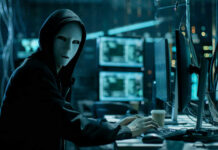 Person in hoodie and mask using computer, multiple screens.
