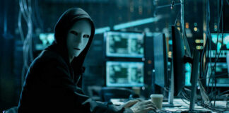 Person in hoodie and mask using computer, multiple screens.