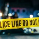 23 Shot As Masked Gunmen Shatter Quiet Night Police line caution tape at a crime scene with blurred figures in the background