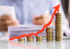 THESE Prices Ignite Economic Alarm Stacked coins with upward red arrow graph in foreground.