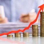 THESE Prices Ignite Economic Alarm Stacked coins with upward red arrow graph in foreground.