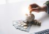A hand placing a coin into a piggy bank surrounded by cash and coins