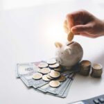 A hand placing a coin into a piggy bank surrounded by cash and coins