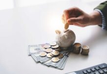 A hand placing a coin into a piggy bank surrounded by cash and coins