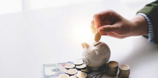 A hand placing a coin into a piggy bank surrounded by cash and coins