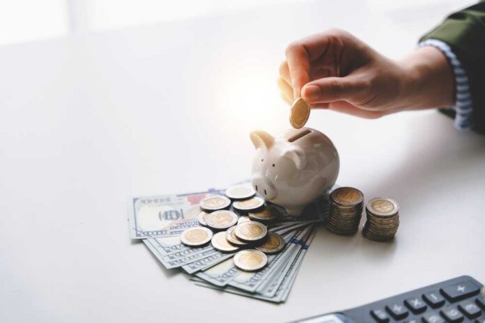 A hand placing a coin into a piggy bank surrounded by cash and coins