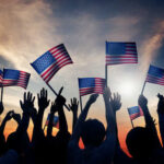 Freedom 250 MELTDOWN — Who Owns Patriotism? Silhouetted crowd holding American flags against a sunset