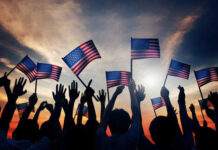 Freedom 250 MELTDOWN — Who Owns Patriotism? Silhouetted crowd holding American flags against a sunset