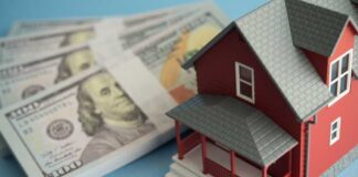 A model red house next to stacks of hundred-dollar bills