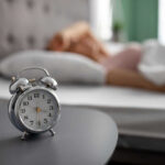 House Roars Yes — Health Risks Loom? An alarm clock on a bedside table with a person sleeping in the background