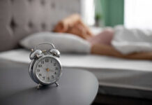 An alarm clock on a bedside table with a person sleeping in the background