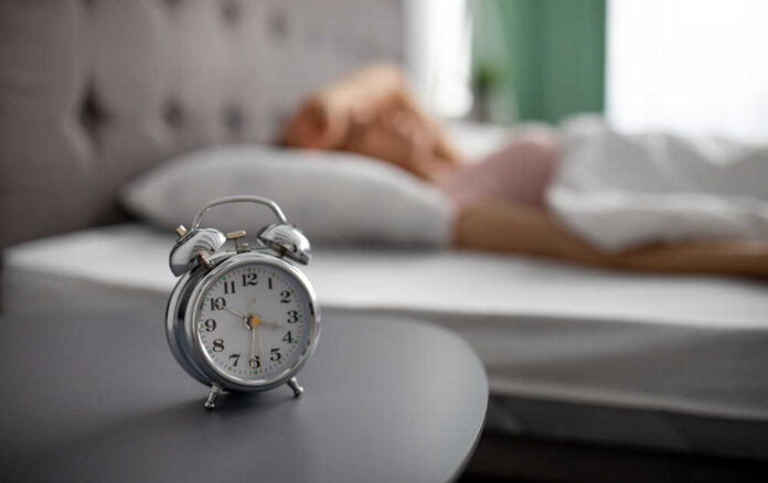 Alarm,Clock,On,Bedside,Table,And,Blurred,View,Of,Millennial An alarm clock on a bedside table with a person sleeping in the background