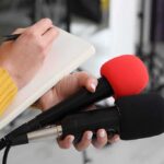 Pop Star Tied To Tesla Body Journalist holding two microphones while writing in a notebook