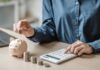 7% Wall Slams THESE Americans Again Person placing a coin into a piggy bank while using a calculator
