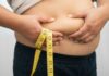 Person pinching belly fat while holding a yellow measuring tape