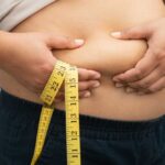 Person pinching belly fat while holding a yellow measuring tape